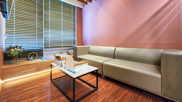 Waiting lounge at TAE Plastic Surgery Clinic with a gray sofa, coffee table, and window blinds.