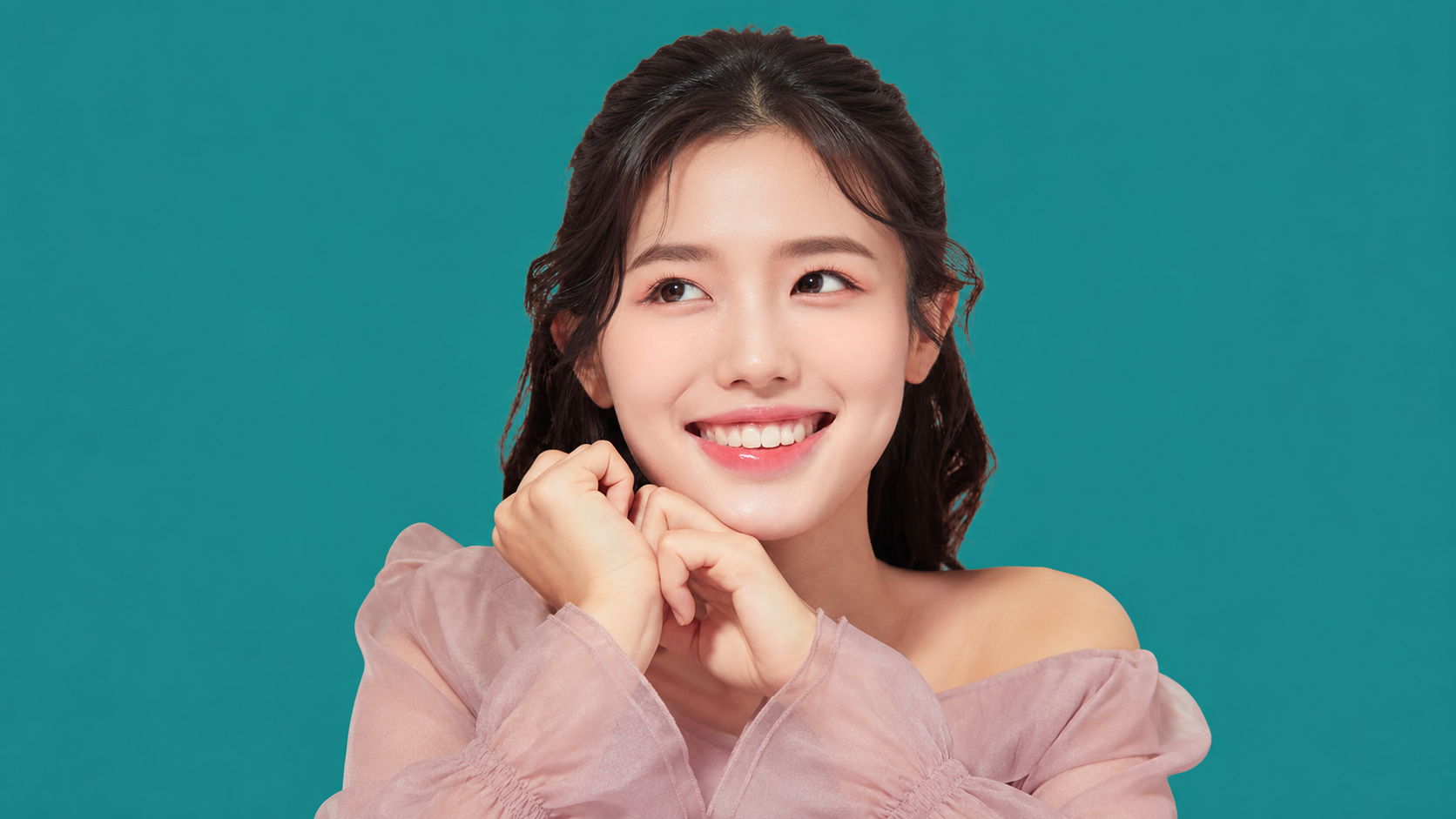 Woman smiling with her hands beneath her chin against a teal background.