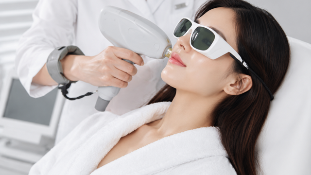 Woman wearing protective goggles receiving a facial light-based treatment with a handheld device.