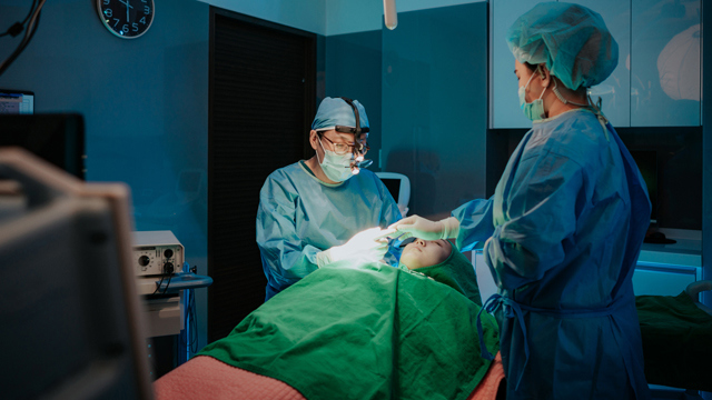 Surgical team wearing masks and surgical gowns performing a procedure on a patient beneath green drapes.
