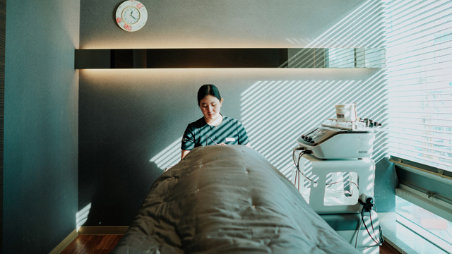 Staff member attending to a reclining patient under a gray blanket beside skincare equipment at TAE Plastic Surgery Clinic.
