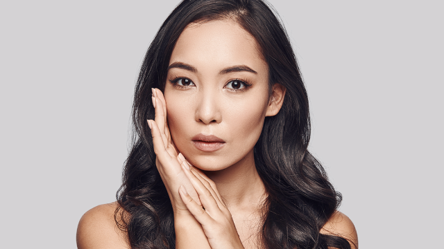 Asian woman touching her smooth face showcasing flawless skin tone and anti-aging dermatology results.