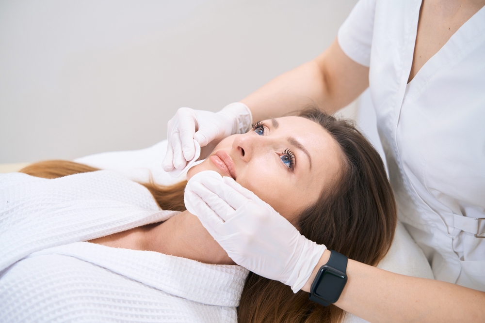 Practitioner wearing white gloves cleansing a reclining woman’s cheeks with cotton pads.