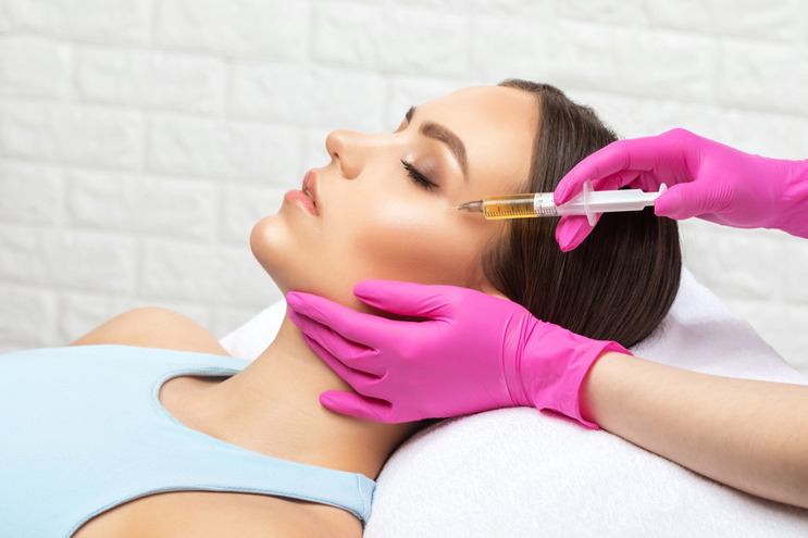 Practitioner wearing pink gloves administering an injection near a reclining woman’s outer eye while supporting her jaw.
