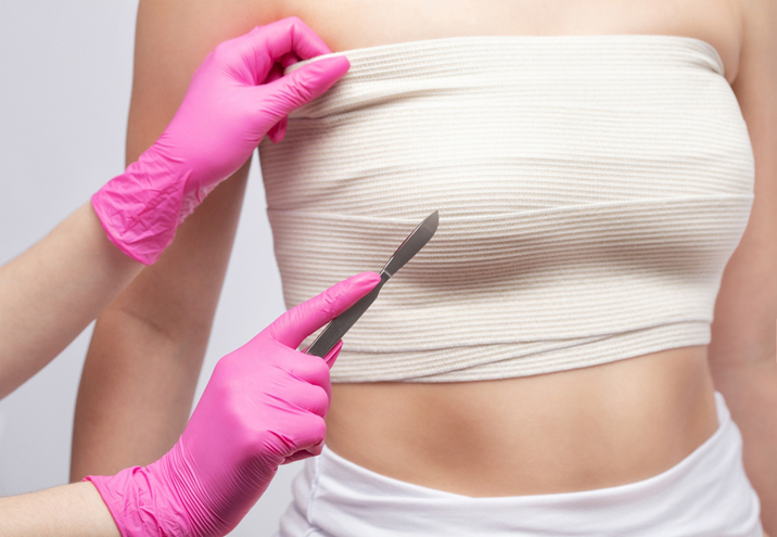 Practitioner wearing pink gloves holding a scalpel beside a woman’s bandaged chest.