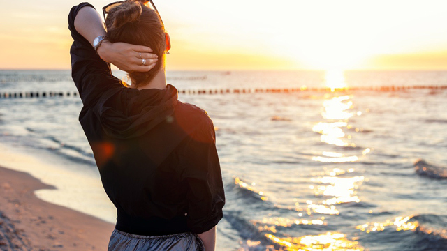 Woman standing on a beach at sunset with one hand resting behind her neck.